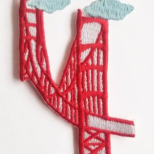 May include: Red embroidered patch of the Golden Gate Bridge with two white clouds above it.