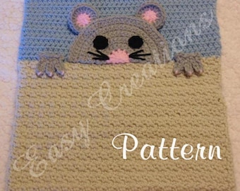 May include: A crocheted mouse-themed blanket pattern. The blanket features a gray mouse head with pink ears and nose, black eyes and whiskers, and gray paws. The blanket is two-toned, with a blue top and tan bottom. The word "Pattern" is written on the blanket.
