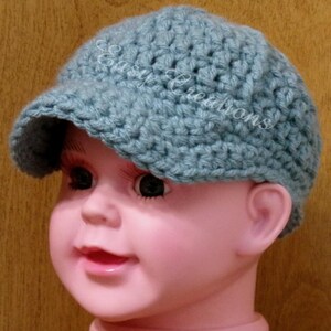 Puede incluir: Un gorro de beb&eacute; de ganchillo azul claro con visera. El gorro est&aacute; en la cabeza de una mu&ntilde;eca beb&eacute;.