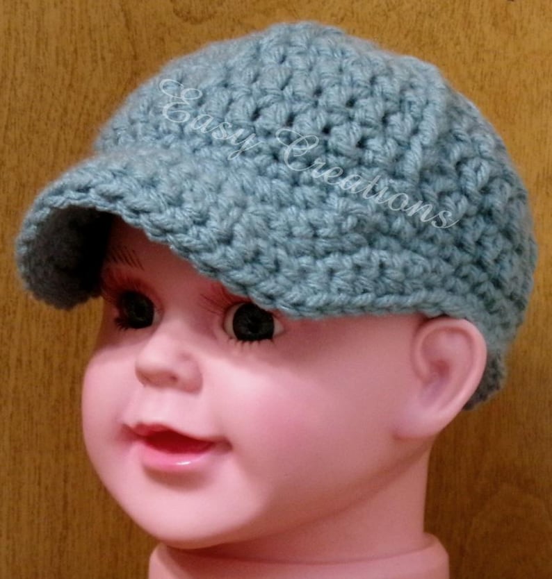 Puede incluir: Un gorro de beb&eacute; de ganchillo azul claro con visera. El gorro est&aacute; en la cabeza de una mu&ntilde;eca beb&eacute;.