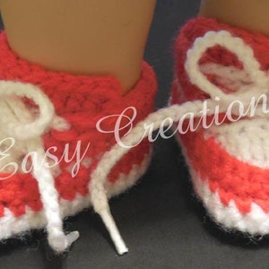 May include: A pair of red and white crocheted baby booties with white laces. The booties have a scalloped edge and are made with a textured stitch.
