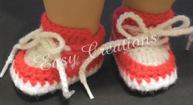 May include: A pair of red and white crocheted baby booties with white laces. The booties have a scalloped edge and are made with a textured stitch.
