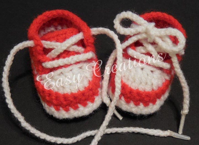 May include: A pair of red and white crocheted baby booties with white laces. The booties have a simple design and are perfect for a newborn.
