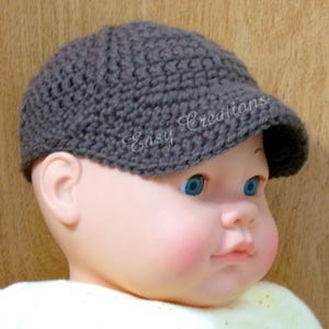 Puede incluir: Un gorro de beb&eacute; de ganchillo gris con visera. El gorro est&aacute; en la cabeza de un beb&eacute;. El beb&eacute; tiene ojos azules y mejillas sonrosadas.