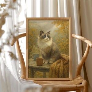 May include: A framed print depicting a fluffy cat with blue eyes, seated on a table with two mugs and a blanket. The cat is grey and white, set against a backdrop of yellow foliage. The frame is a light brown colour.
