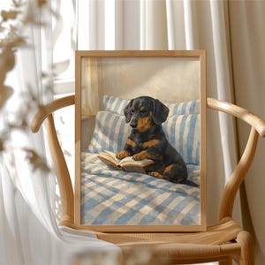 Op de afbeelding: Een ingelijste prent van een zwarte en bruine teckel die een boek leest op een bed met blauw-wit gestreepte kussens en een geruite sprei. De lijst is van licht hout en wordt op een houten stoel gepresenteerd.