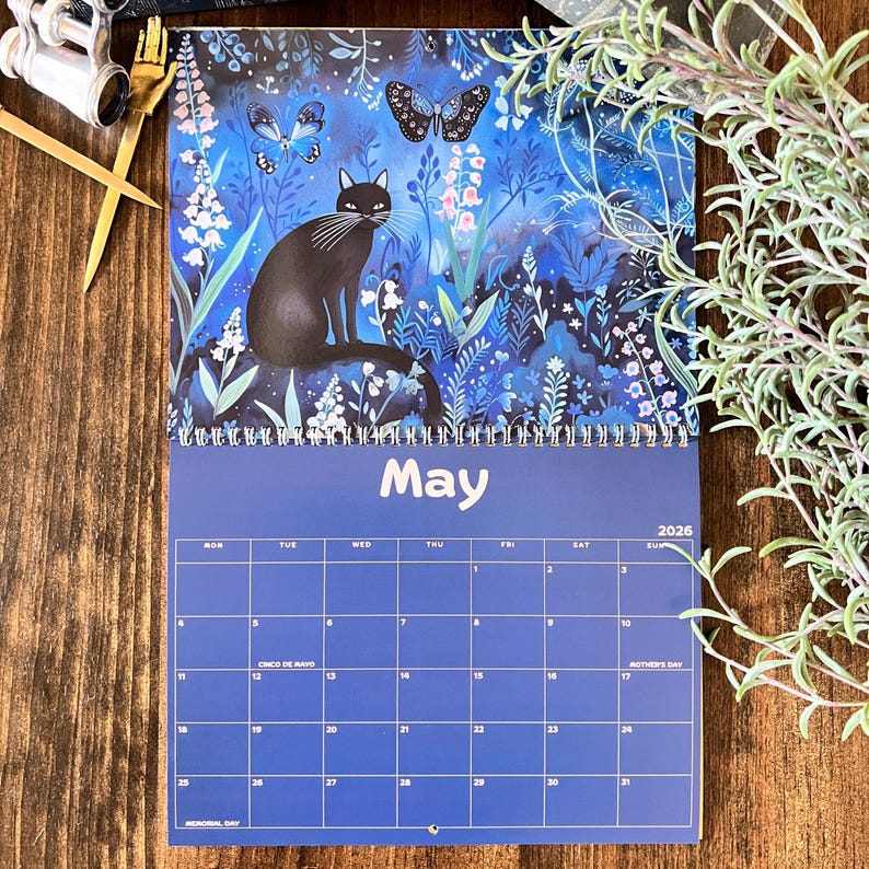 May include: A spiral-bound calendar for May 2026, featuring a black cat illustration set against a blue floral background with butterflies. The calendar has a blue grid with dates and holidays.