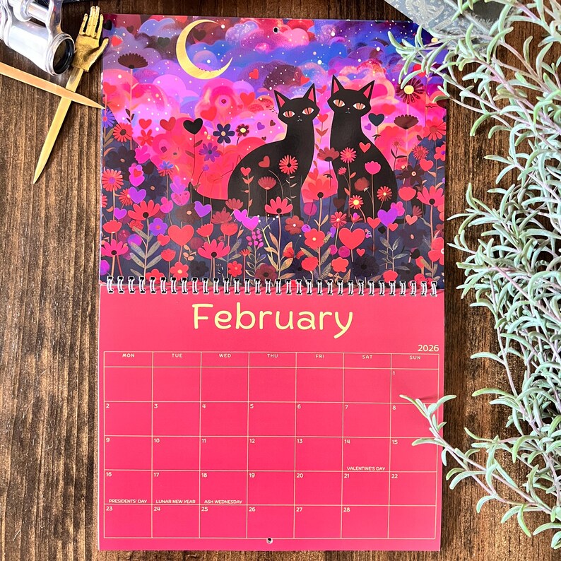 May include: A 2026 wall calendar featuring a vibrant illustration of two black cats amidst a field of flowers and hearts under a crescent moon. The calendar is predominantly pink with the month "February" displayed prominently.