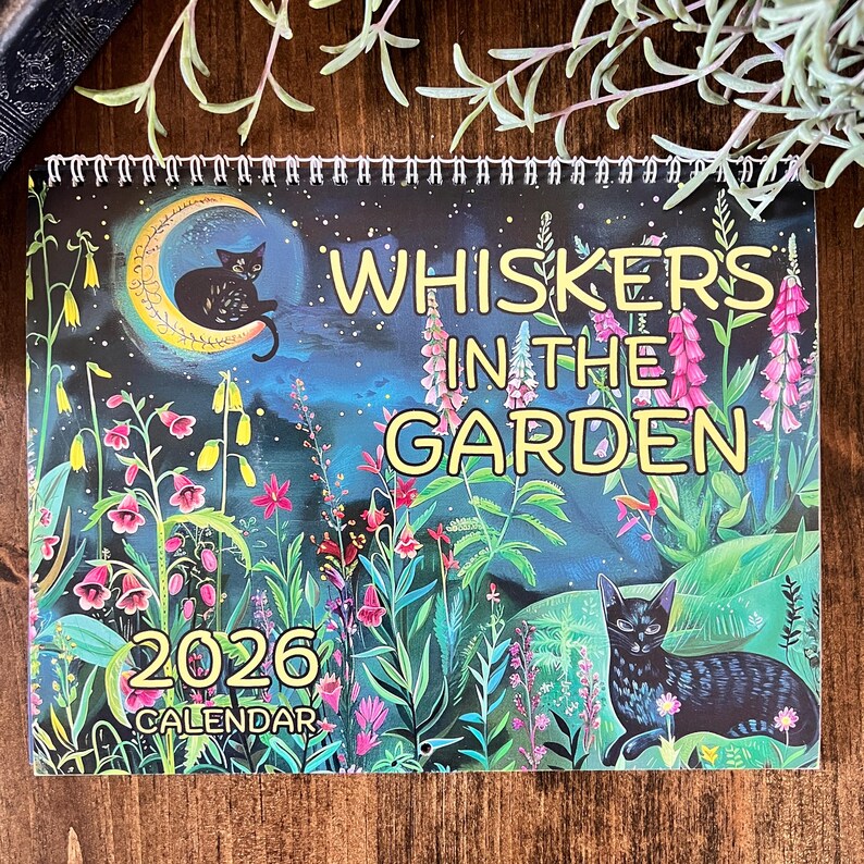 May include: A spiral-bound 2026 calendar titled "Whiskers in the Garden." The cover features a black cat on a crescent moon, and another cat among colorful flowers. The background is a dark blue night sky.