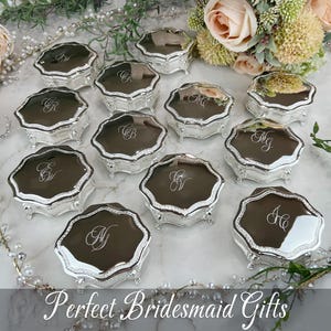 May include: A collection of small, silver-toned, octagonal trinket boxes with ornate feet and reflective lids. Each lid features a unique, engraved script monogram. The boxes are arranged on a marble surface with floral accents and pearl strands.