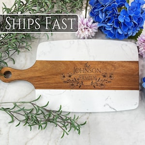 May include: A rectangular serving board featuring a central horizontal strip of brown wood flanked by white marble sections. The wood center is engraved with the text the JOHNSON FAMILY and floral illustrations. It includes a handle with a hole.