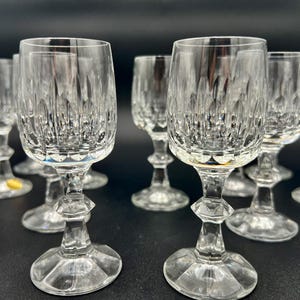 May include: A collection of clear crystal wine glasses. Each glass features a detailed, cut-glass design on the bowl and a faceted stem. The glasses are arranged on a dark surface, reflecting light and creating a sparkling effect.