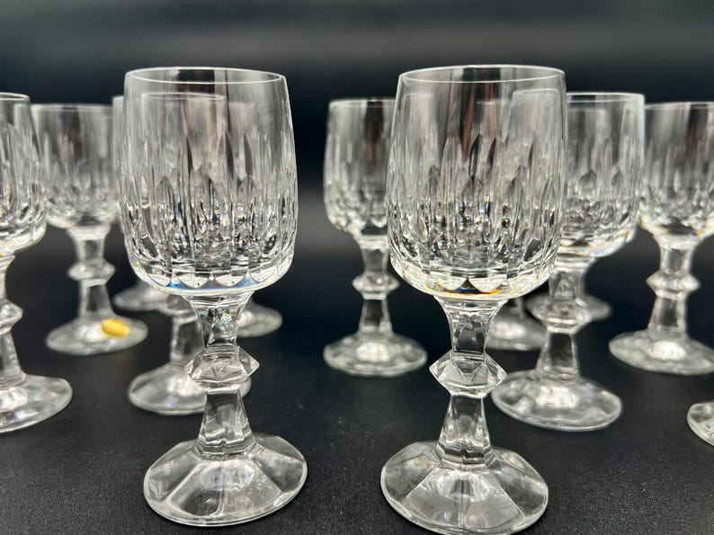 May include: A collection of clear crystal wine glasses. Each glass features a detailed, cut-glass design on the bowl and a faceted stem. The glasses are arranged on a dark surface, reflecting light and creating a sparkling effect.