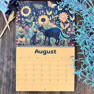 May include: A spiral-bound wall calendar for 2026 featuring a whimsical illustration of black cats in a floral garden. The month of August is displayed in large, bold letters. The calendar has a yellow background with a weekly grid.
