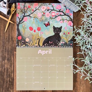 May include: A 2026 wall calendar featuring a black cat in a garden scene with pink flowers and a butterfly. The word "April" is printed in light pink. The calendar has a spiral binding and a grid of dates.