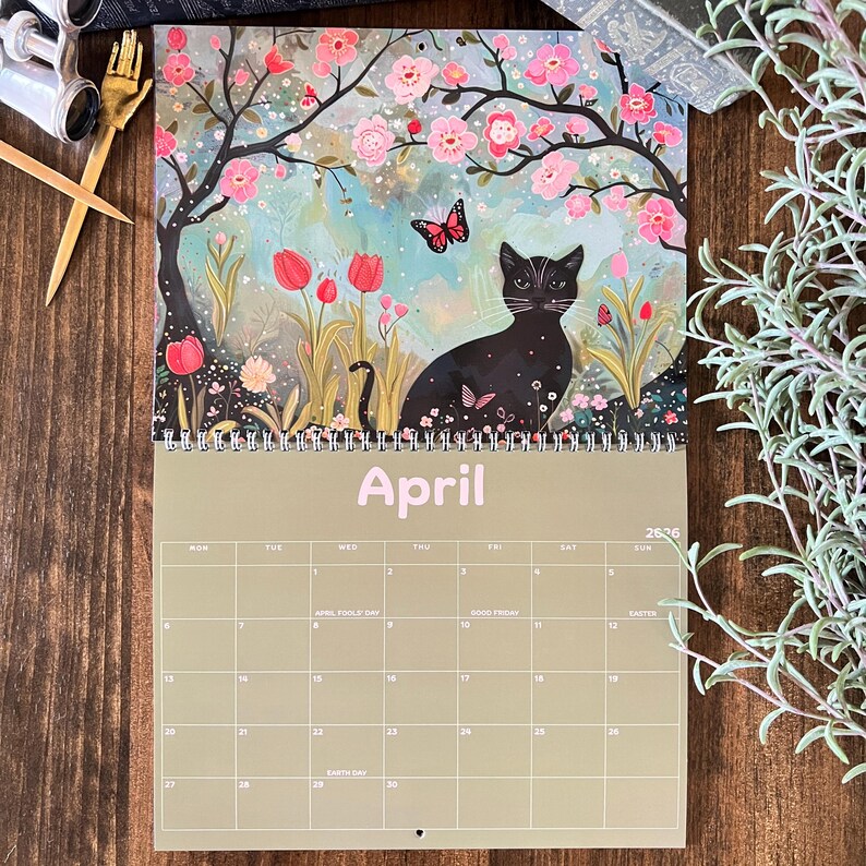 May include: A 2026 wall calendar featuring a black cat in a garden scene with pink flowers and a butterfly. The word "April" is printed in light pink. The calendar has a spiral binding and a grid of dates.