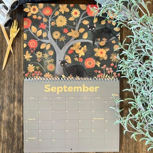 May include: A 2026 calendar featuring a whimsical illustration of black cats in a tree with orange and yellow flowers. The month of September is displayed in gold lettering. The calendar is on a wooden surface.