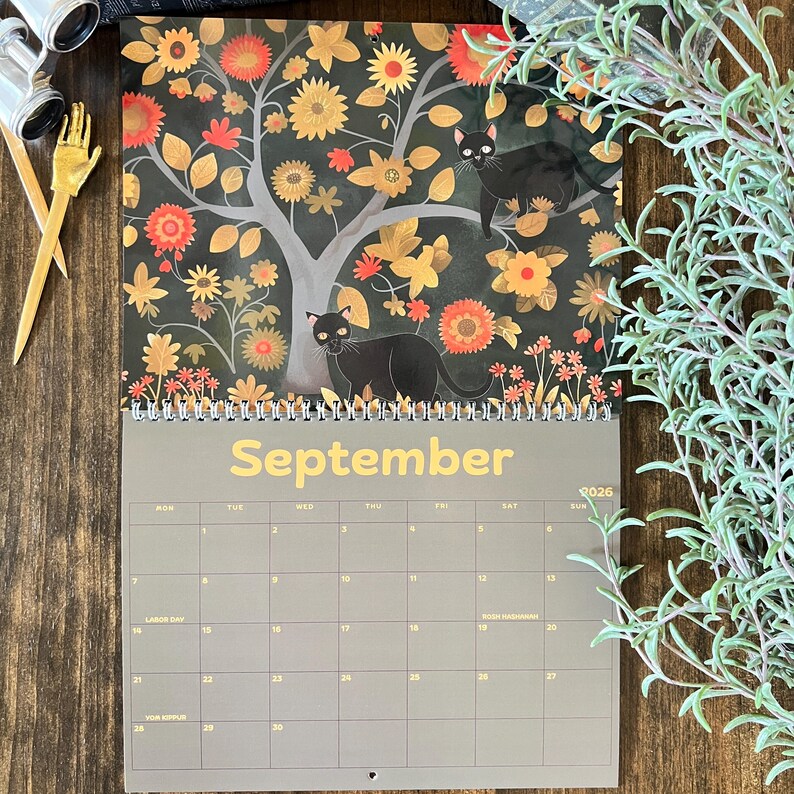 May include: A 2026 calendar featuring a whimsical illustration of black cats in a tree with orange and yellow flowers. The month of September is displayed in gold lettering. The calendar is on a wooden surface.