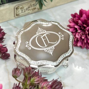 May include: A silver-toned metal trinket box with a scalloped edge and a reflective taupe lid. The lid features an engraved white oval frame containing the stylized monogram letters CFL. The box has ornate silver detailing along the sides.