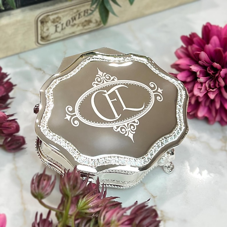 May include: A silver-toned metal trinket box with a scalloped edge and a reflective taupe lid. The lid features an engraved white oval frame containing the stylized monogram letters CFL. The box has ornate silver detailing along the sides.