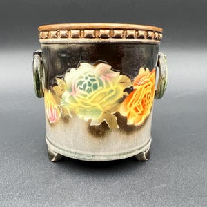 May include: A ceramic planter with a dark brown top and a light brown base. The planter features a floral design with yellow, orange, and pink roses. It has two green handles and sits on three small feet.