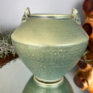 May include: A green ceramic vase with a textured surface and frog-shaped handles. The vase has a wide, rounded body and a narrow base. The color is a muted green with darker accents around the rim and handles. The vase is reflected in the surface below.