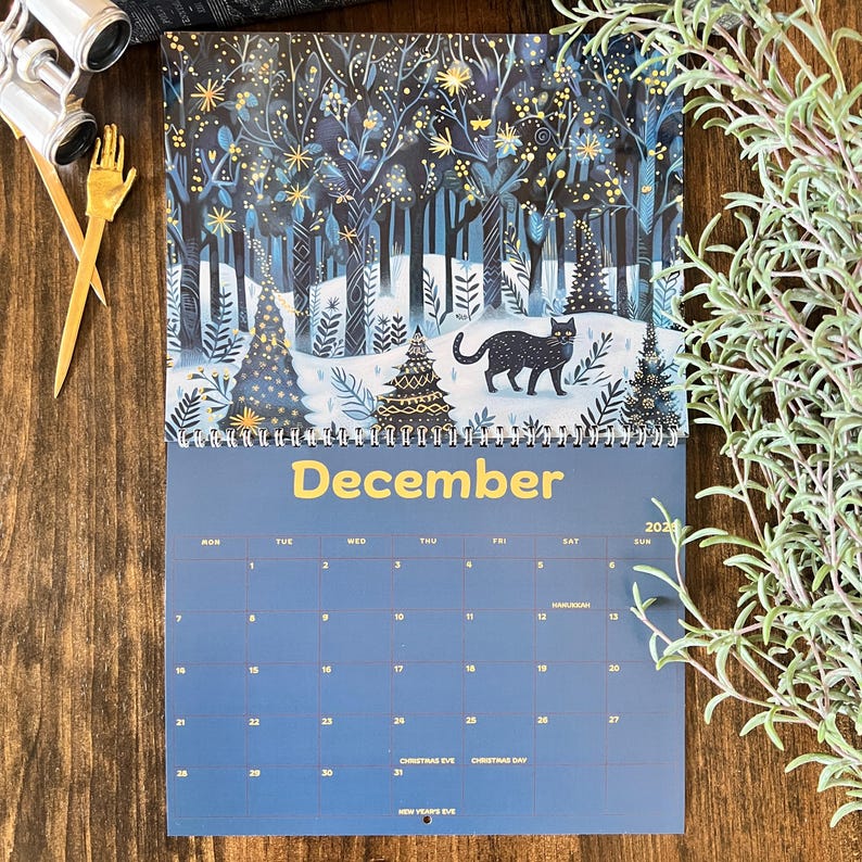 May include: A December 2024 wall calendar featuring a winter forest scene with a black cat. The top portion displays a snowy forest with trees and a black cat. The word "December" is in yellow. The calendar grid is blue.