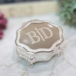 May include: A silver-toned trinket box with a scalloped edge and a smooth, taupe-colored lid featuring the engraved letters B|D. The base of the box is supported by four ornate, detailed feet, and the sides are decorated with a textured, metallic pattern.