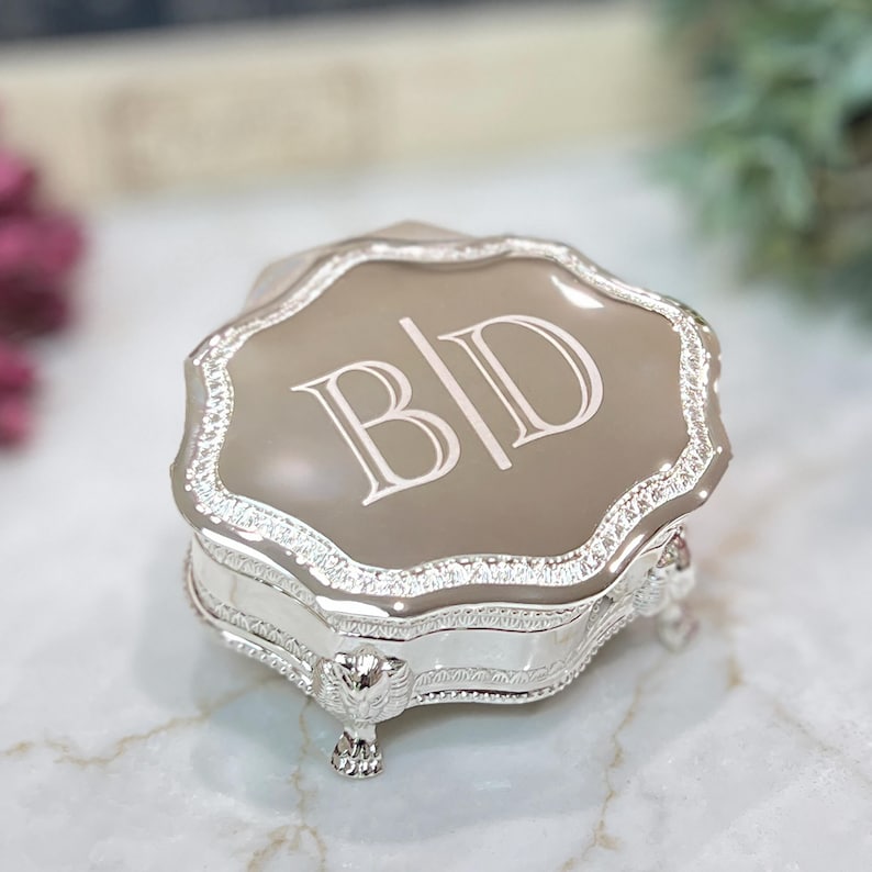 May include: A silver-toned trinket box with a scalloped edge and a smooth, taupe-colored lid featuring the engraved letters B|D. The base of the box is supported by four ornate, detailed feet, and the sides are decorated with a textured, metallic pattern.