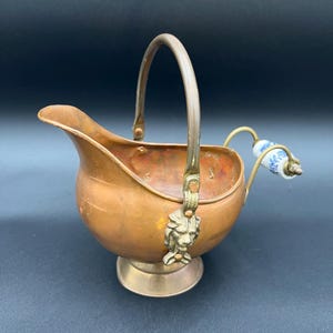 May include: A copper-colored metal sugar bowl with a brass base and handle. The handle is attached with a lion's head design. The small handle on the side has a white and blue floral design.