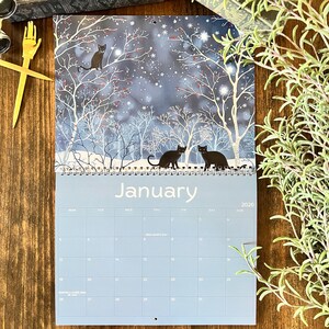 May include: A 2026 wall calendar featuring a winter scene with black cats. The artwork depicts a snowy forest with bare trees and a starry night sky. The word "January" is printed at the top of the calendar.