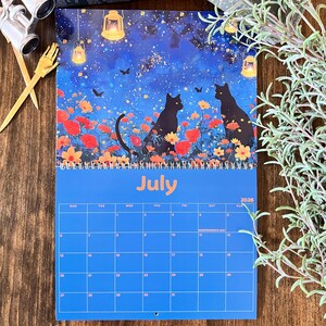 May include: A July 2026 calendar featuring a whimsical illustration of two black cats in a flower field under a starry night sky. The calendar has a blue background with the month and year prominently displayed.