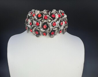 May include: A silver-toned floral collar necklace with red gemstone centers. The necklace is displayed on a white mannequin against a dark background. The flowers are detailed and three-dimensional.