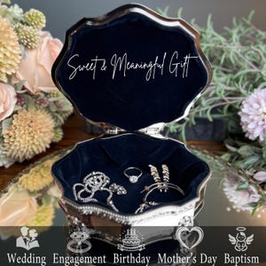 May include: A silver-toned metal jewelry box with a scalloped edge and dark blue velvet lining. The interior lid features the white text Sweet & Meaningful Gift. Inside, a silver chain, a ring, and hoop earrings are displayed on the velvet.