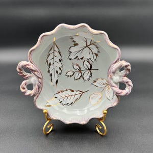 May include: A white ceramic serving dish with a scalloped edge and two handles. The dish features gold leaf designs and a gold-colored stand. The handles and rim have a pinkish-purple accent. The dish is likely vintage.