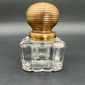 May include: Clear glass inkwell with a brass-colored, ribbed ball-shaped lid. The inkwell has a square base with rounded corners. The lid is attached to a brass-colored ring. The inkwell is empty.