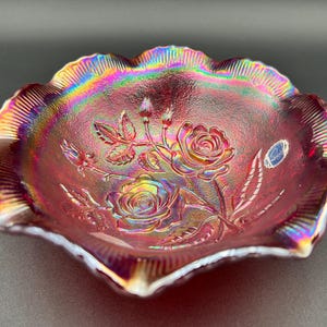 May include: A ruby-red iridescent glass bowl with a ruffled edge. The bowl features an embossed rose design with leaves and buds. The rim has a rainbow sheen, and the bowl is likely vintage.