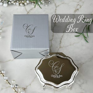 May include: A silver square box and a metallic silver octagonal ring box with ornate feet. Both items feature a custom engraving of the monogram CS, the names Chris & Sara, and the date 6.27.2026. The ring box has a reflective lid surface.