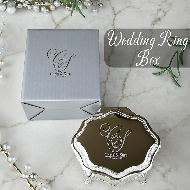 May include: A silver square box and a metallic silver octagonal ring box with ornate feet. Both items feature a custom engraving of the monogram CS, the names Chris & Sara, and the date 6.27.2026. The ring box has a reflective lid surface.