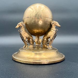 May include: A vintage brass decorative object featuring a spherical top with a hinged lid, supported by a pedestal base. Two dog figures with chain details flank the sphere. The piece has a polished, aged appearance.