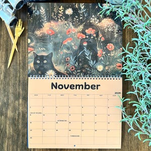 May include: A spiral-bound calendar for November 2026, featuring a whimsical illustration of two black cats amidst a floral landscape. The calendar has a beige background with the days of the week and dates clearly marked.