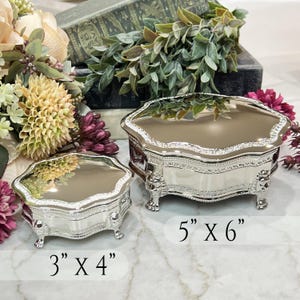 May include: Two silver-toned metal trinket boxes with reflective lids and ornate lion-shaped feet. The smaller box measures 3 inches by 4 inches, and the larger box measures 5 inches by 6 inches. Both containers feature decorative embossed edges.