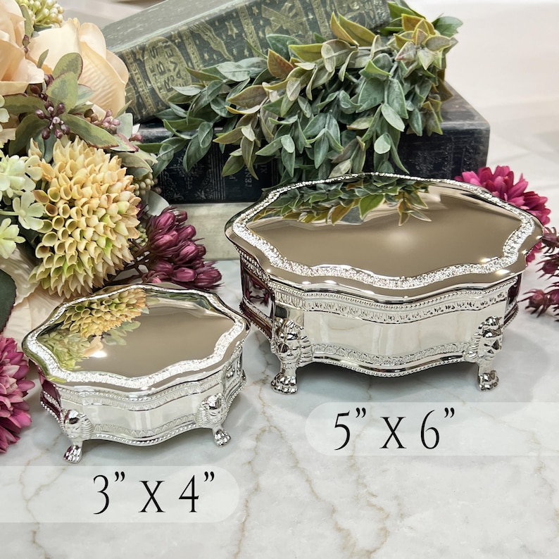 May include: Two silver-toned metal trinket boxes with reflective lids and ornate lion-shaped feet. The smaller box measures 3 inches by 4 inches, and the larger box measures 5 inches by 6 inches. Both containers feature decorative embossed edges.