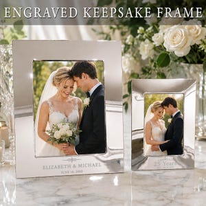 May include: Two rectangular silver-toned metal picture frames with polished surfaces. The larger frame is engraved with "ELIZABETH & MICHAEL" and "JUNE 14, 2000". The smaller frame is engraved with "25 Years" and "EST. 06.14.2000". Both frames hold wedding photos.