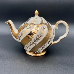 May include: A white and gold teapot with a lid and handle. The teapot features gold stripes and leaf designs. The spout and handle are white. The teapot sits on a gold base and has a gold finial on the lid.