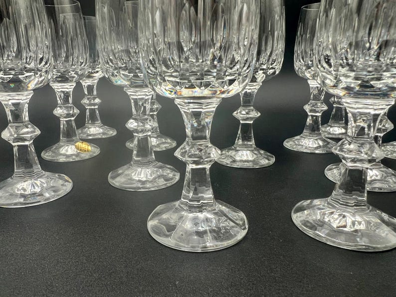 May include: A collection of clear crystal wine glasses. The glasses have a detailed, textured design on the bowl and stem. The stems have a decorative, bulbous shape. The glasses are arranged on a dark surface.