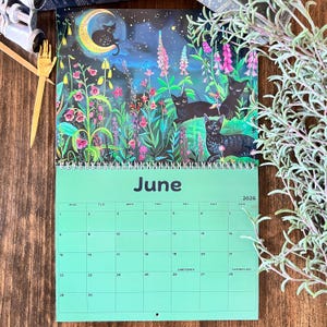 May include: A 2026 wall calendar featuring a whimsical illustration of black cats in a moonlit garden. The calendar page for June is displayed, with a green background and dates. The artwork includes vibrant colors and floral elements.