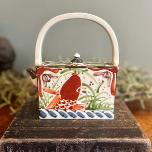 May include: A square ceramic teapot with a white handle and spout. The teapot is decorated with a red fish design, green plants, and blue waves. The lid is topped with a small black and white finial. The teapot is sitting on a dark wooden surface.