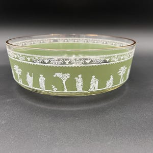May include: A clear glass bowl with a gold rim and a light green interior. The exterior features a white decorative band with a classical scene of figures and trees. The bowl is likely vintage and suitable for serving.