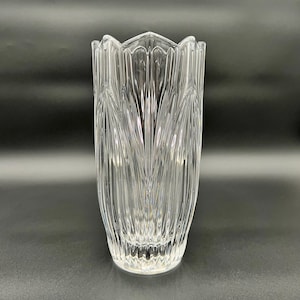 May include: A clear glass vase with a fluted design. The vase has a tall, cylindrical shape with a scalloped rim. Vertical lines run the length of the vase, creating a textured appearance. The vase is empty and sits against a dark background.